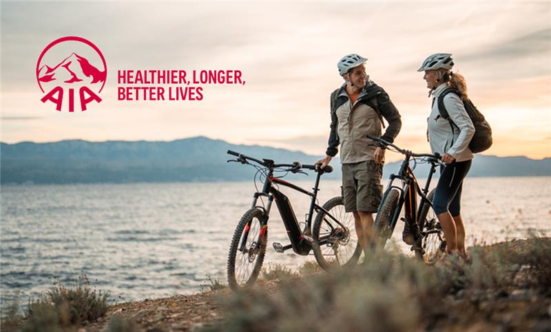 Two cyclists stand with their bikes on grass near a body of water, with mountains and a cloudy sky in the background during sunrise or sunset.